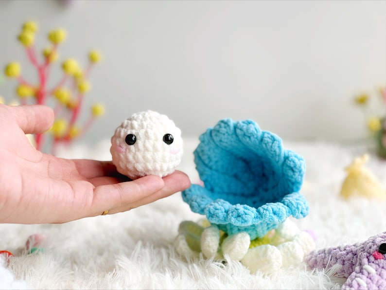 May include: A white crocheted plush toy with black eyes and a pink mouth, sitting inside a blue crocheted shell with white crocheted accents.