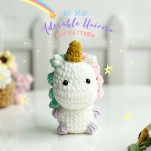 May include: A white crocheted unicorn with a gold horn, light blue and pink accents, and black eyes. The unicorn is sitting on a white surface with a white picket fence in the background. The text "NO SEW Adorable Unicorn PDF PATTERN" is above the unicorn.