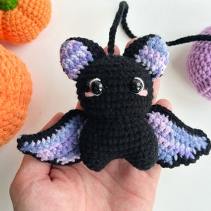 May include: A crocheted black bat with purple and blue wings. The bat has large black eyes and pink cheeks.