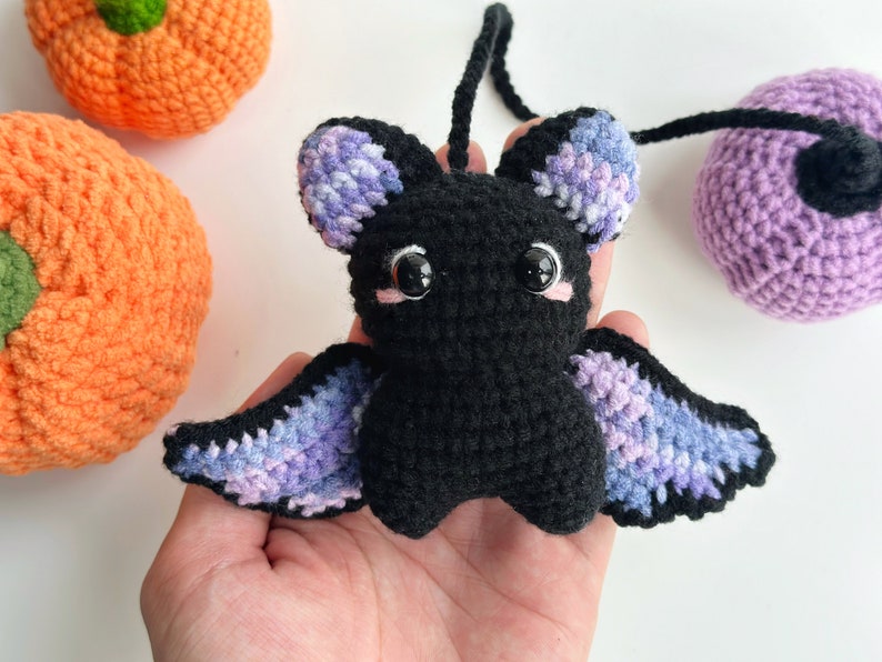 May include: A crocheted black bat with purple and blue wings. The bat has large black eyes and pink cheeks.