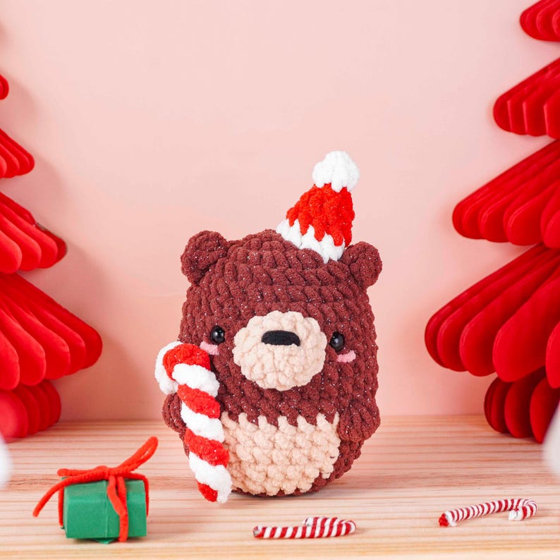 May include: A handmade brown crochet bear wearing a red and white Santa hat, holding a candy cane. The bear has a light tan muzzle and belly. The background is a light pink with red paper Christmas trees.