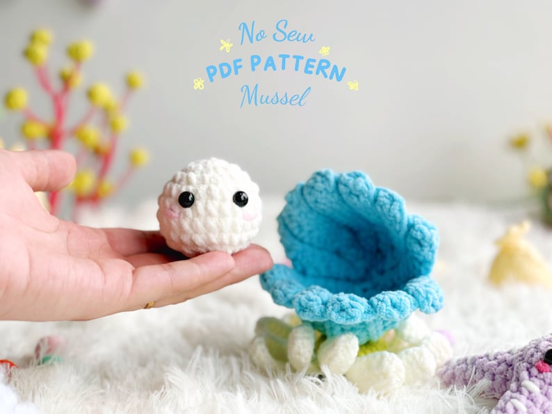 May include: A white crocheted amigurumi mussel with black eyes and pink cheeks, sitting inside a blue crocheted shell. The text "No Sew PDF PATTERN Mussel" is written above the amigurumi.