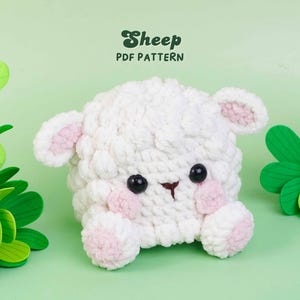 May include: A white crocheted sheep plush with pink accents on the ears and cheeks, featuring black plastic eyes and a brown stitched nose. The text Sheep PDF PATTERN is printed in dark green above the plush on a light green background.