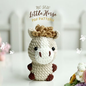 May include: A crocheted stuffed horse toy with a brown mane and a white face. The toy is sitting on a white surface with a white picket fence in the background. The text "NO SEW Little Horse PDF PATTERN" is written above the toy.