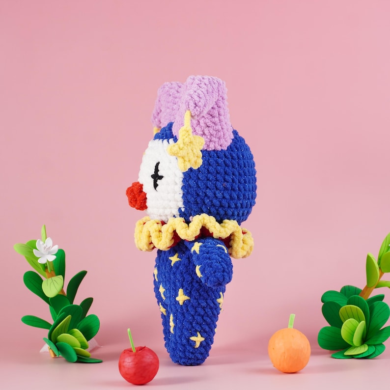 May include: A handmade crochet clown doll with a blue body adorned with yellow stars. The doll has a white face, red nose, and a yellow ruffled collar. The clown's hat is lavender, and it has a yellow star on its head. The background is pink.