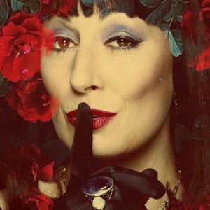 May include: A woman with dark hair and red lipstick is wearing a crown of red roses and a black glove with a large, dark blue gemstone ring. She is holding her finger to her lips, as if to tell someone to be quiet.