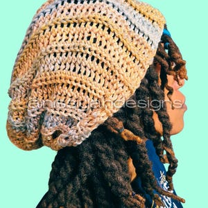 May include: A crocheted beanie in shades of tan, beige, and light gray. The hat has a loose, open weave design. The image shows the side profile of a person wearing the hat, with long dreadlocks visible.