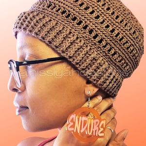 May include: A brown crocheted hat with a textured design. The hat is worn with black-framed glasses and orange earrings that read "ENDURE". The earrings are circular and have a pink outline.