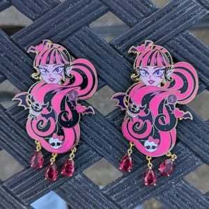 Op de afbeelding: Twee roze en zwarte emaillepins met een cartoon-vampiermeisje met vleermuisvleugels en een vleermuis. De pins hebben gouden accenten en hangende roze edelstenen.