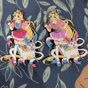 Könnte beinhalten: Zwei Emaille-Anstecknadeln mit einem blonden Charakter in einem rosa Kleid und einem Sylveon. Die Anstecknadeln haben einen goldenen Umriss und sind vor einem dunkelblauen Hintergrund mit floralem Muster platziert. Der Charakter hält einen Pokéball.
