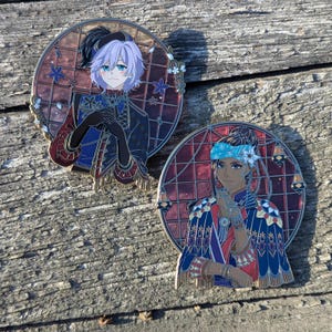 Può includere: Due spille rotonde in smalto con personaggi in stile anime. Una spilla raffigura un personaggio con capelli lavanda e un cappello nero, l'altra un personaggio con capelli scuri e una fascia blu. Entrambe le spille hanno dettagli intricati e un bordo metallico.