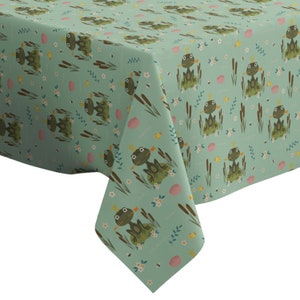 Op de afbeelding: Een groene tafelkleed met een herhalend patroon van kikkers met kronen, zittend op waterlelies, omringd door riet en bloemen.