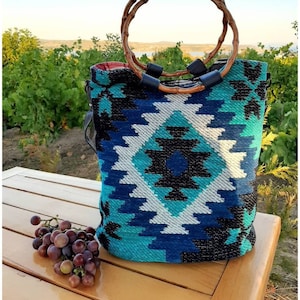 May include: A blue and white patterned tote bag with a woven design. The bag has two brown wooden handles with black leather accents. The bag is sitting on a wooden table with a bunch of grapes in front of it.