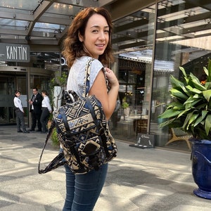 May include: A woman wearing a gray shirt and blue jeans is standing in front of a building. She is carrying a black and brown backpack with a geometric pattern. The backpack has a large pocket on the front and a smaller pocket on the side. The woman is looking over her shoulder at the camera.