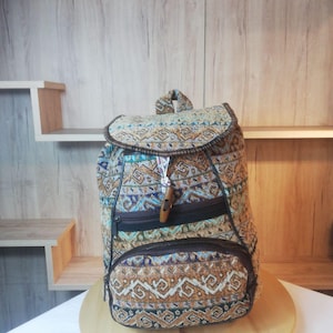 Könnte beinhalten: Ein Rucksack aus gewebtem Stoff mit geometrischem Muster in Braun-, Beige-, Blau- und Cremetönen. Die Tasche verfügt über eine Klappe mit Holzknebelverschluss, eine Reißverschlusstasche auf der Vorderseite und dunkelbraune Kantenbesätze.