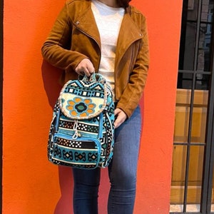 Puede incluir: Una mochila con un vibrante patrón tejido en tonos turquesa, negro, blanco y naranja. La bolsa tiene una solapa superior redondeada con un prominente motivo de flor naranja, un cierre de cordón y un bolsillo frontal con cremallera y diseños geométricos.