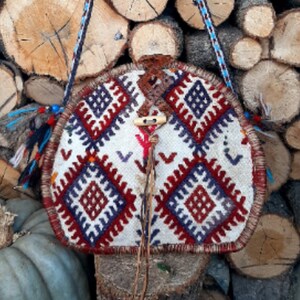 May include: A round, white, red, and blue patterned bag with a brown leather strap. The bag is hanging from a wooden log background.
