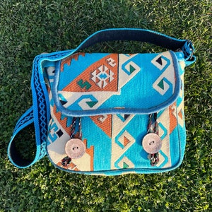 May include: A blue and orange patterned crossbody bag with a black strap and two large buttons. The bag is made of woven fabric and has a geometric design.
