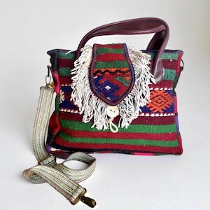 May include: A colorful woven handbag with a fringed flap and leather handles. The bag features a striped pattern in red, green, and blue, with a decorative flap secured by a button. It includes a detachable, patterned shoulder strap.