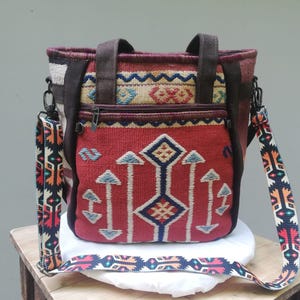 Op de afbeelding: Een tote bag met een geweven geometrisch patroon in rood, crème en blauw. De tas heeft donkerbruine stoffen handvatten, een voorvak met ritssluiting en een afneembare schouderriem met een kleurrijk, herhalend tribaal patroon.