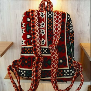 May include: A red, black, white and green patterned backpack with thick, braided straps. The backpack is made of woven fabric and has a traditional design.