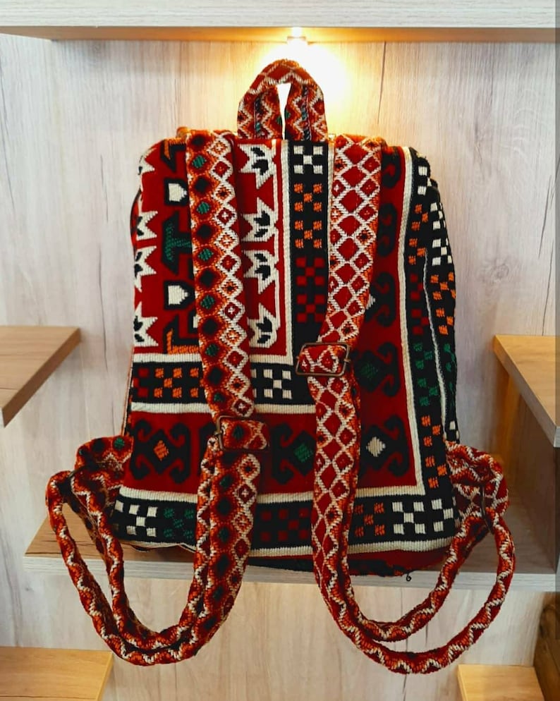 May include: A red, black, white and green patterned backpack with thick, braided straps. The backpack is made of woven fabric and has a traditional design.