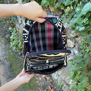 May include: A black and white patterned backpack with a plaid lining. The backpack has a zippered pocket on the front and a zippered closure at the top.
