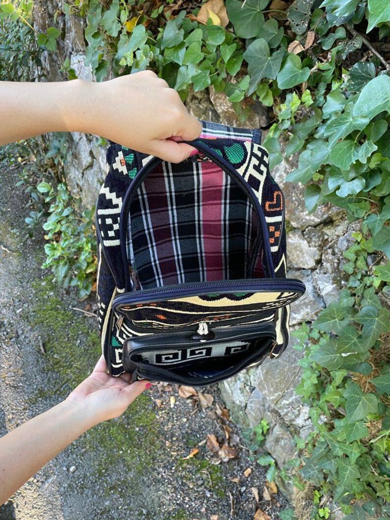 May include: A black and white patterned backpack with a plaid lining. The backpack has a zippered pocket on the front and a zippered closure at the top.