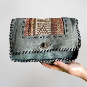 May include: A gray leather clutch with a woven fabric flap and a metal clasp. The clutch has a decorative stitched border.