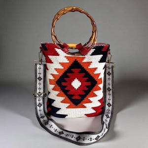 May include: A woven tote bag with a geometric pattern in red, orange, black, and white. It has a bamboo handle and a detachable shoulder strap with a repeating diamond design. The bag is rectangular in shape.