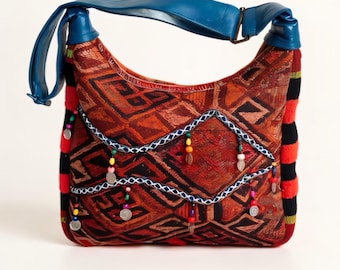 Op de afbeelding: Een hobo-tas met een stoffen body in een rood en oranje geometrisch patroon en een blauwe schouderband. De voorzijde is versierd met een witte gestikte golvende rand, kleine hangende zilverkleurige metalen munten en kleurrijke kralen. De zijkanten hebben zwarte en rode strepen.