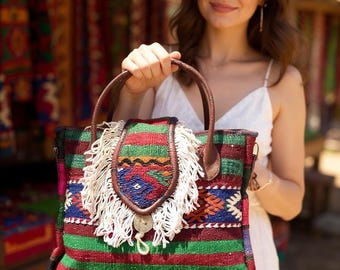 May include: A colorful, patterned tote bag with brown leather handles and a decorative flap with white fringe. The bag features a mix of red, green, and blue woven stripes and geometric designs, with a button closure.