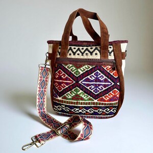 May include: A colorful tote bag with a geometric pattern in red, purple, green, and white. The bag has brown suede handles and trim, a zippered pocket, and a patterned shoulder strap. The bag is designed for everyday use.