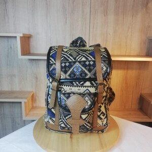 May include: A backpack featuring a woven fabric with a geometric pattern in shades of blue, black, white, and tan. The bag includes tan faux leather straps with metal buckles, a front pocket with a flap closure, and black trim along the edges.