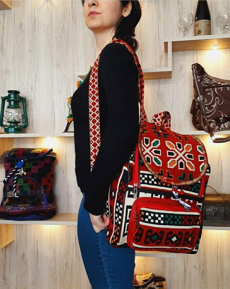 May include: A red, black and white woven backpack with a geometric pattern. The backpack has a large main compartment and a smaller front pocket. The straps are red and white.