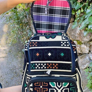 May include: A black and white patterned backpack with green and orange accents. The backpack has a flap that opens to reveal a plaid lining in pink, black, and purple.