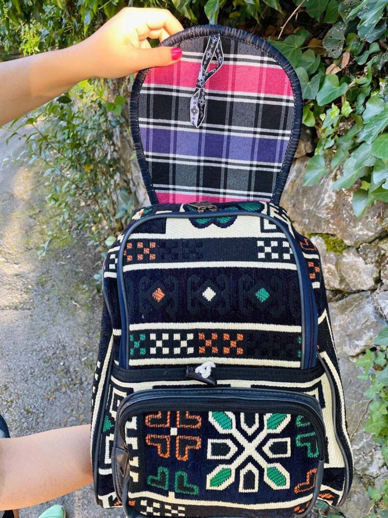 May include: A black and white patterned backpack with green and orange accents. The backpack has a flap that opens to reveal a plaid lining in pink, black, and purple.