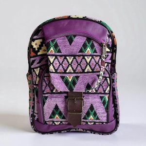 May include: Purple backpack with a geometric pattern in shades of purple, green, orange, and yellow. The backpack has a brown leather buckle and a zipped pocket on the front.