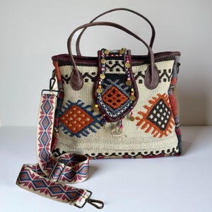 Könnte beinhalten: Beige Tragetasche mit farbenfrohem geometrischen Muster in Orange, Blau und Schwarz. Sie hat braune Ledergriffe und einen abnehmbaren gemusterten Riemen. Die Tasche hat dekorative Verzierungen und einen Klappenverschluss.