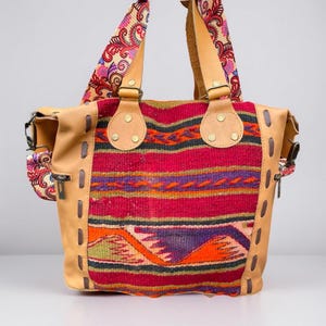 May include: A tan leather tote bag with a colorful woven fabric panel featuring geometric patterns in red, orange, purple, green, and black. The bag has two top handles and a detachable shoulder strap.