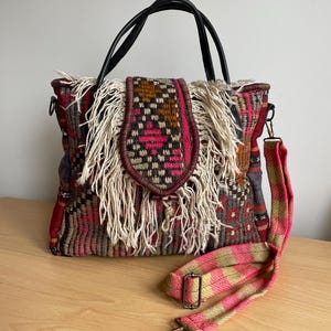 May include: A woven tote bag with a flap closure and fringe. The bag features a black leather handle and a detachable pink and tan striped shoulder strap. The bag's woven pattern includes red, brown, and pink hues.