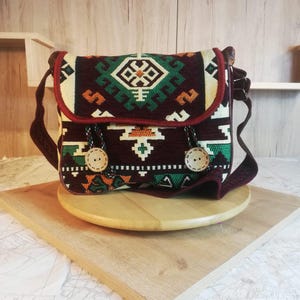 May include: A rectangular shoulder bag featuring a dark maroon base with a woven geometric pattern in green, cream, and orange. The flap is secured by two large, light-colored wooden buttons with dark cord loops and a maroon fabric trim.