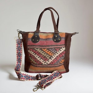 May include: A brown tote bag with a colorful woven front panel featuring geometric patterns in red, orange, and beige. It has brown suede sides, leather handles, and a detachable patterned strap with metal clasps. The bag is designed for carrying items.