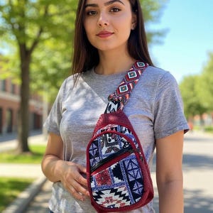 May include: A crossbody sling bag featuring a dark red textured exterior and a front panel with a multi-colored geometric pattern in blue, white, black, and red. The bag includes a matching patterned adjustable shoulder strap and a black zipper.