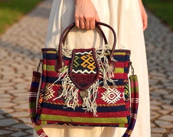 May include: A colorful tote bag with a patterned design. The bag features a mix of red, green, blue, and white stripes, with a decorative flap and fringe. It has brown leather handles and a matching shoulder strap.