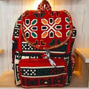 May include: A red, black, green and white patterned backpack with a brown woven handle. The backpack has a smaller red, black, green and white patterned pouch attached to the front.