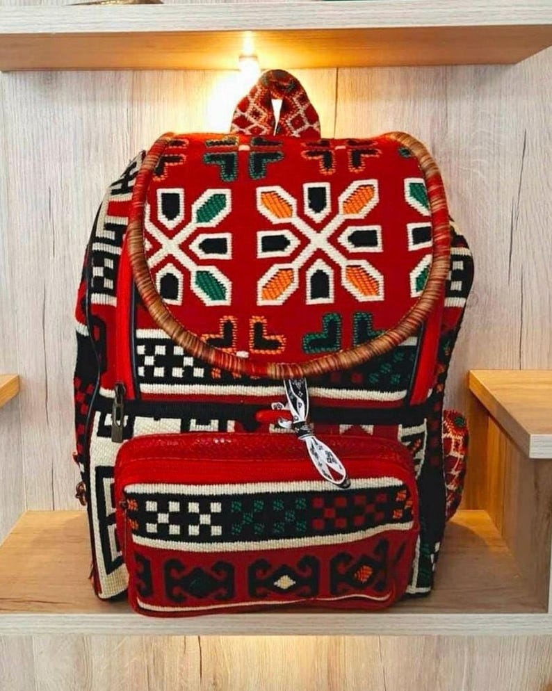 May include: A red, black, green and white patterned backpack with a brown woven handle. The backpack has a smaller red, black, green and white patterned pouch attached to the front.