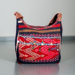 Könnte beinhalten: Eine rote, schwarze und braune gewebte Tragetasche mit bunten Perlenfransen und silbernen Akzenten. Die Tasche hat einen Lederriemen.