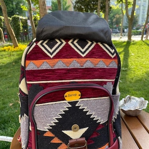 May include: A burgundy backpack with a black and white geometric pattern. The backpack has a flap with a buckle closure and a small pocket on the front. The name "Serap" is written on a gold tag on the front of the backpack.