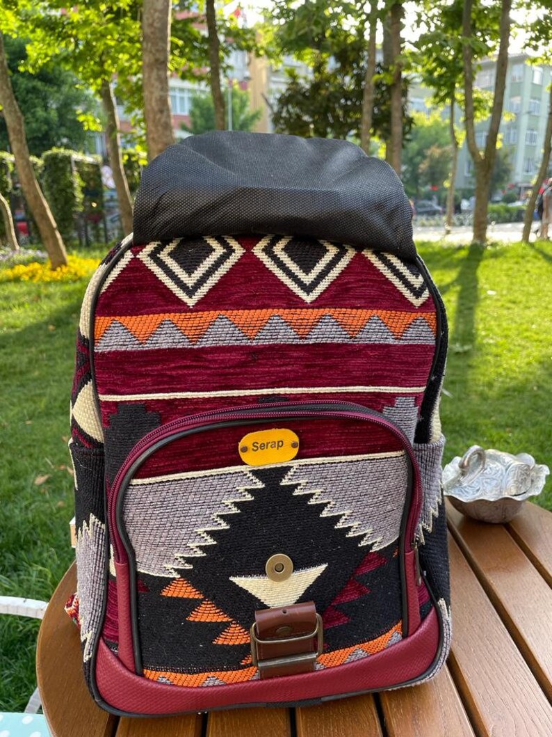 May include: A burgundy backpack with a black and white geometric pattern. The backpack has a flap with a buckle closure and a small pocket on the front. The name "Serap" is written on a gold tag on the front of the backpack.